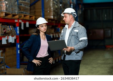 Caucasian adult female and male supervisors wearing helmets discussing warehouse operations while reviewing logistics data on laptop during stock inspection for delivery scheduling and management