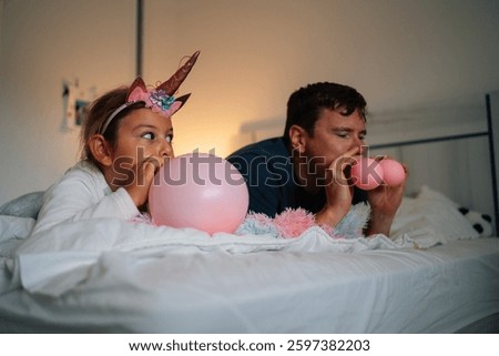 Caucasian adult male and young hispanic girl blowing pink balloons on bed.