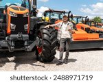 Caucasian adult man with digital tablet in hand stands next to construction or agricultural tractor at a machinery dealer. Ag equipment dealer or supplier.