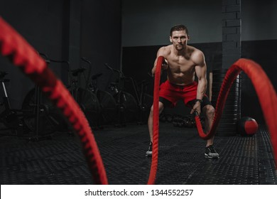 Caucasian athlete doing battle ropes exercise at the crossfit gym. Man wear red shorts training with rope. The sport motivation concept.