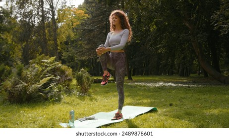 Caucasian athlete sportswoman woman standing on one leg balancing holding pressing knee to chest sport stretching yoga balance stretch flexibility workout yoga on mat in park at nature girl exercise - Powered by Shutterstock