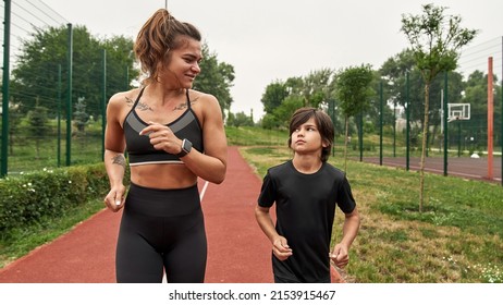 Caucasian Athletic Mother And Son Jogging Or Running On Running Track Outdoors. Adult Woman With Tattoos And Boy Looking At Each Other. Modern Healthy And Sports Lifestyle. Family Spend Time Together