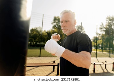 Caucasian attractive handsome determined active senior man, wearing black t-shirt and elastic bands on wrists, boxing with punching bag in outdoor boxing ring on sunny summer day. Martial arts. Sport  - Powered by Shutterstock