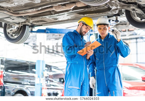 Caucasian automotive mechanic man writing on\
maintenance document clipboard and checking under car in auto\
repair garage, assistant pointing flash light, transport business\
and after service\
concept