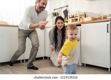 Caucasian Baby Boy Child Learn To Walk With Parents Support In House. Happy Family, Mother And Father Helping Young Toddler Son Taking First Step Walk On Floor To Develop Skill In Living Room At Home.