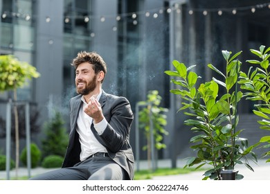 Caucasian Bearded Business Man Smoking Cannabis Outdoors Sitting On City Park Bench On Urban Street Background. Male Employee Businessman. Office Worker In Suit Relieves Stress A Marijuana Outside