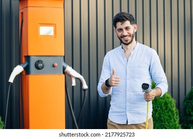 Caucasian bearded hipster man standing near electric charging station looking at camera and showing thumb up. Male holds a charging cable in his hands. Eco-friendly modern green transport.