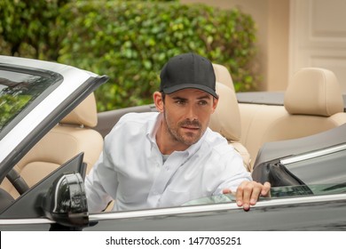 A caucasian bearded man steps out of his convertible looking away to the right. Holding the car door open, he remains focused while wearing a black baseball cap and buttondown white shirt.