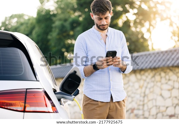 Caucasian bearded man\
using smart phone and waiting power supply connect to electric\
vehicles for charging the battery in car. Plug charging an Electric\
car from charging\
station.