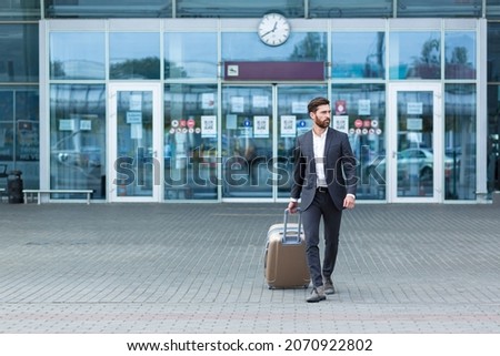 Caucasian bearded tourist businessman walks public transport building with luggage on urban background a city street. Man in formal suit trip. Business traveler pulling suitcase in airport terminal