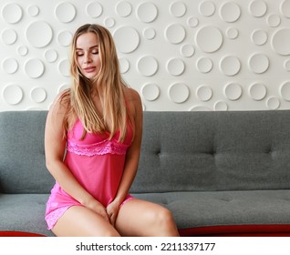 Caucasian Beautiful Blonde Hair Hot Female Fashion Model In Pink Sexy Lingerie Underwear Pajamas Kneeling Sensual Seductive Temptation Posing On Gray Cozy Sofa Couch In Living Room At Apartment.