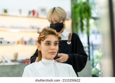 Caucasian Beautiful Woman Customer Using Mobile Phone Checking Modern Types Of Hairstyles And Consult With Experts Hairstylist. Hairdresser Wearing Facemask To Prevent From Coronavirus In Beauty Salon