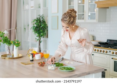 Caucasian beautiful woman preparing fresh salad ripe strawberries and arugula leaves, diet breakfast luxurious girl in a beautiful light dress with makeup and hairstyle. Happy in the kitchen at home