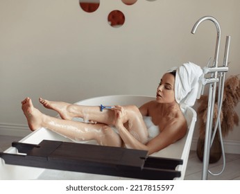 Caucasian Beautiful Woman Shaving Legs Using Blue Razor With Foam Taking Bath In Modern Bathroom. Happy Female Removing Body Hair While Taking Bath In Tub At Home. High Angle View. Beauty Routine