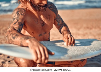 Caucasian beefy bearded man with tattoos in swimming trunks is waxing a surfboard near the sea. Sports and active recreation. - Powered by Shutterstock