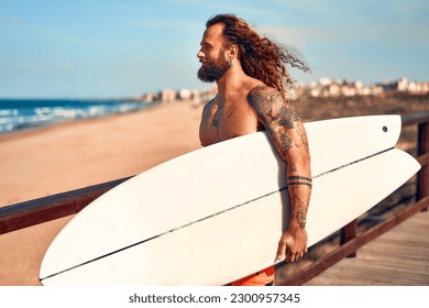 Caucasian beefy bearded man with tattoos in swimming trunks holding a surfboard walks along a wooden bridge to the sea. Sports and active recreation. - Powered by Shutterstock