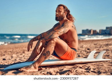 Caucasian beefy surfer bearded man with tattoos and long hair in swimming trunks with a surfboard near the sea. Sports and active recreation. - Powered by Shutterstock