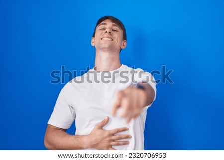 Caucasian blond man standing over blue background laughing at you, pointing finger to the camera with hand over body, shame expression 