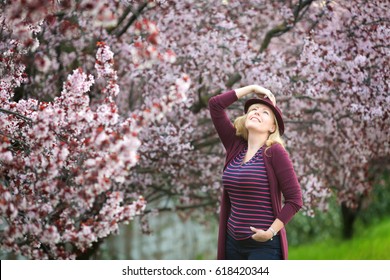 Caucasian Blond Woman With Long Hair In Purple Fedora Hat Near Blossoming Tree