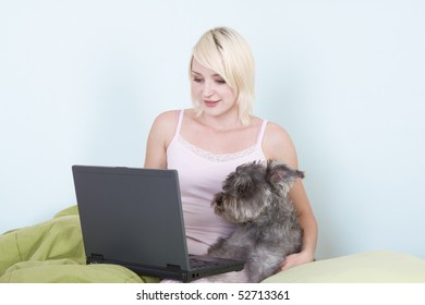 Caucasian Blond Young Woman Sitting On Bed Working On Portable Notebook Pc Computer Together With Dog Of Schnauzer Breed