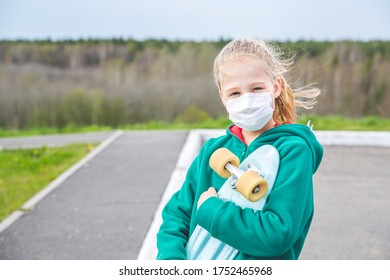 Caucasian Blonde Girl In A Green Coat Holds A Blue Skateboard In Her Hands. Girl In A Medical Mask.The Wind Is Blowing Hair.The Forest In The Background. Solo Outdoor Activities.Covid-19 Coronavirus.