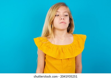 Caucasian blonde kid girl wearing yellow T-shirt against blue wall making grimace and crazy face, screaming out of control, funny lunatic expressing freedom and wild.