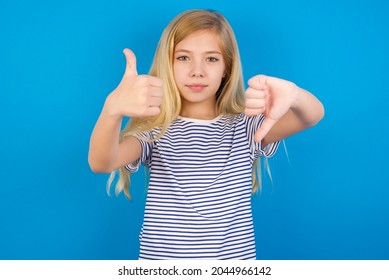 Caucasian Blonde Kid Girl Wearing Stripped T-shirt Against Blue Wall Showing Thumbs Up And Thumbs Down, Difficult Choose Concept