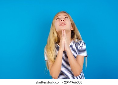 Caucasian Blonde Kid Girl Wearing Stripped T-shirt Against Blue Wall Begging And Praying With Hands Together With Hope Expression On Face Very Emotional And Worried. Please God