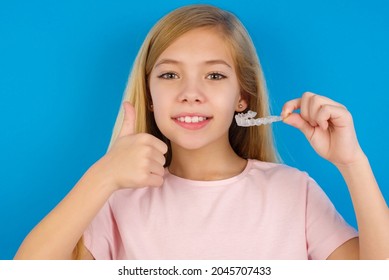 Caucasian Blonde Kid Girl Wearing Pink T-shirt Against Blue Wall Holding An Invisible Braces Aligner And Rising Thumb Up, Recommending This New Treatment. Dental Healthcare Concept.