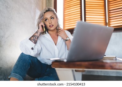 Caucasian blonde woman with digital netbook technology using wireless connection on mobile phone for calling and talking indoors, skilled freelancer making consultancy conversation via cellphone