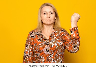 Caucasian blonde woman in her 40s feeling serious, strong and rebellious, raise her fist, protest or fight for revolution against flat yellow wall. - Shutterstock ID 2309998179