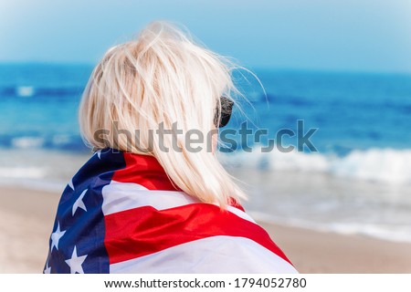 Caucasian blonde woman holding american flag leaning against back and looking at sea