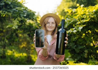 Caucasian blonde woman vintner relaxing in the vineyards in summer season, shows smartphone and bottle of red wine to camera. Girl in hat and an apron holding red wine bottle outdoors. Outdoor farmer - Powered by Shutterstock