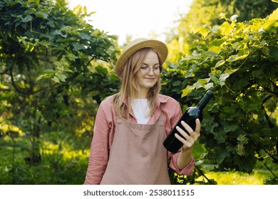 Caucasian blonde woman vintner relaxing in the vineyards in summer season. Girl in hat and an apron holding red wine bottle outdoors. Outdoor farmer countryside style. High quality photo - Powered by Shutterstock