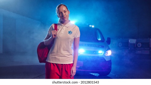 Caucasian Blonde Young Female Paramedic In Uniform Getting Ready To Work At Night Shift. Ambulance Car On Background. Good Looking Woman Doctor Outdoor With Backpack. Portrait. Call 911. Paramedic