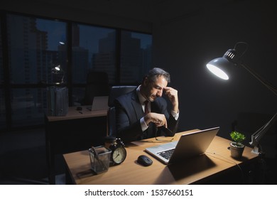 Caucasian boss working late sitting on desk in office at night. Business man feeling tired and stress for overload job hold glasses and hand on nose