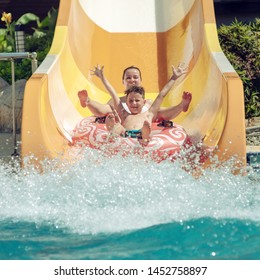 Caucasian boy and mom gliding down slide in waterpark. They enjoy the fun and holding hands wide open.