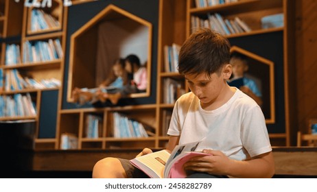 Caucasian boy reading a book while group of smart students sitting at library. Attractive child studying, learning from novel or textbook while children talking, chitchat about education. Erudition. - Powered by Shutterstock