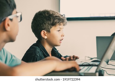 Caucasian boy reading task aloud during lesson. Classmates and pupils sitting at table in school and using laptops for study. Knowledge, childhood, communication and digital education concept - Powered by Shutterstock