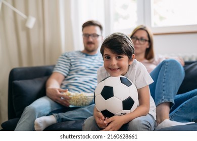 Caucasian Boy Sitting On A Sofa And Holding Soccer Ball While Watching Game With His Parents