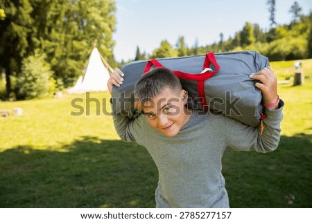 CAUCASIAN BOY SMILING WHILE HOLDING THE PACKED TENT STANDING AT THE CAMPSITE, HAPPY TEENAGER LOVING LIVING WITH NATURE, SUMMER VACATION CONCEPT, CAMPING IN THE GREEN FOREST NATURE