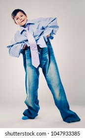 Caucasian Boy Wearing His Dad's Shirt, Jeans And Tie On Light Background. He Is Wearing Big Adult Size Clothes Which Are Too Big For Him.