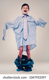 Caucasian Boy Wearing His Dad's Shirt, Jeans And Tie On Light Background. He Is Wearing Big Adult Size Clothes Which Are Too Big For Him.