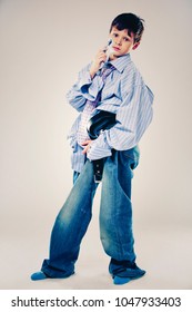 Caucasian Boy Wearing His Dad's Shirt, Jeans And Tie On Light Background. He Is Wearing Big Adult Size Clothes Which Are Too Big For Him.