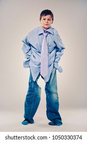 Caucasian Boy Wearing His Dad's Shirt, Jeans And Tie On Light Background. He Is Wearing Big Adult Size Clothes Which Are Too Big For Him.