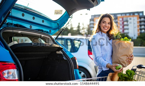 Caucasian brunette going holding paper bags with\
food products. Young woman putting package with groceries and\
vegetables into car trunk. Attractive caucasian female shopping in\
mall or grocery store