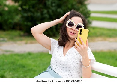 Caucasian brunette woman in trendy round sunglasses. Attractive girl in white dress and modern sunwear makes selfie sitting on bench in city park on cloudy summer day.
