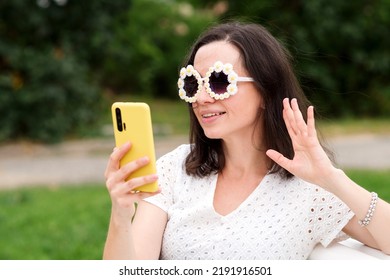 Caucasian brunette woman in trendy sunwear makes selfie sitting on bench in city park. Girl in white dress and optical round sunglasses communicates via video call in cell mobile phone outdoors. 