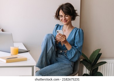 Caucasian brunette woman using phone . European cute smiling woman holding smartphone, wear blue blouse sitting at home with laptop. Freelance concept 