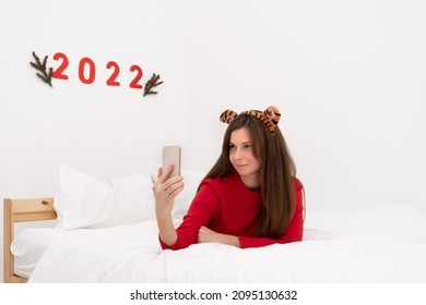 Caucasian Brunette Woman,long Hair Wear Cozy Red Sweater,tiger Ears,ly On White Bed Taking Selfie Photo With Smartphone On Background Of 2022 On Wall.New Year Holiday On Eastern Calendar.Copy Space.